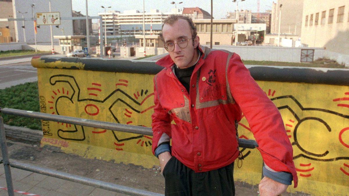 Why is an AI altered Keith Haring painting sparking outrage? Pictured here: Haring stands in front of part of the Berlin Wall that he painted in Berlin - 1986