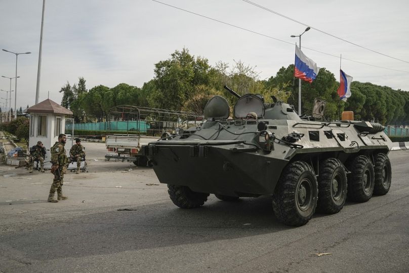 Un véhicule blindé russe passe devant les combattants syriens qui gardent l'aéroport de Lakatia, dans la ville de Khmeimim, au sud-est de la ville de Lattaquié, en Syrie, le lundi 16 décembre 2024.