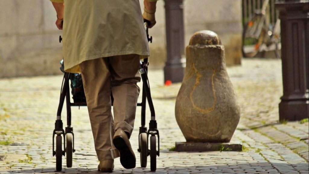 An elderly man walks using a walker.