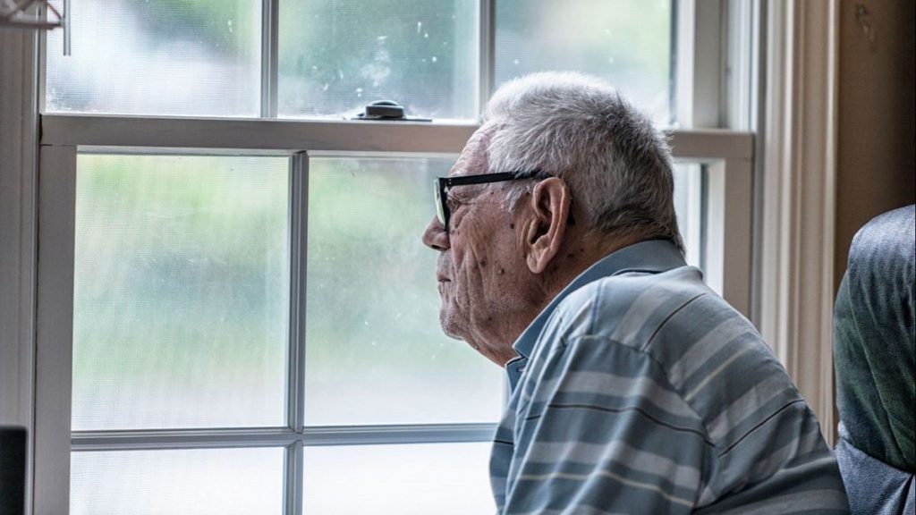 An elderly man looks out the window.