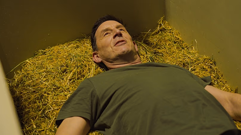 Chris Cooper-Hayes allongé dans un récipient rempli de luzerne, de paille et de copeaux de bois à Return Home.