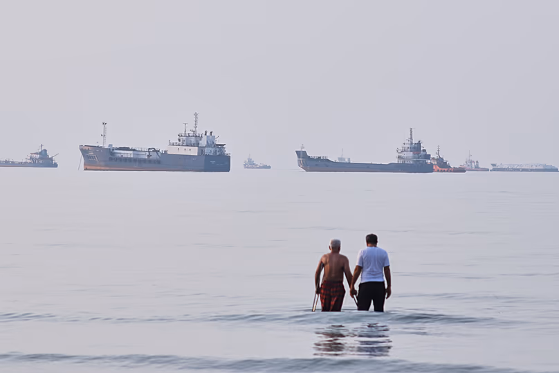 Deux hommes pataugent dans les eaux du détroit d'Ormuz avec des navires ancrés en arrière-plan, au large de Bandar Abbas, en Iran, le dimanche 12 juillet 2026.