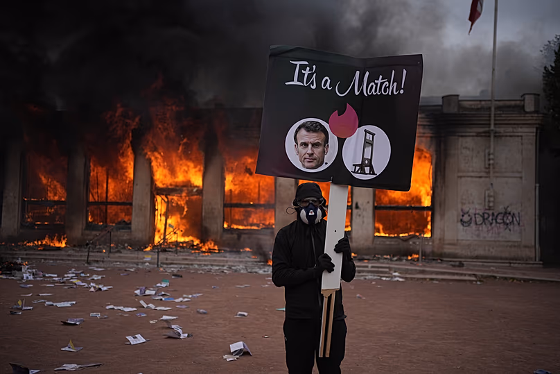 Un manifestant tient un panneau avec une photo du président français Emmanuel Macron devant un office du tourisme en feu lors d'une manifestation à Lyon, France, le samedi 17 octobre 2026.